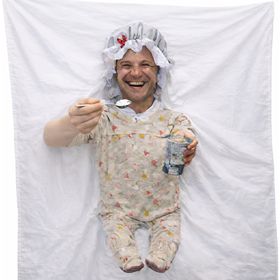 Mann im Babykostüm lacht während eines Hochzeitsspiels, vor dekorierter Kulisse mit Lichtern und festlicher Atmosphäre.