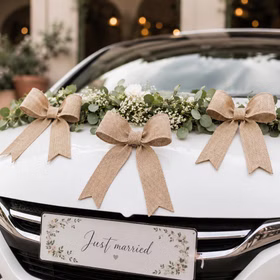 Rustikale Jute-Schleifen als Hochzeitsauto-Deko an weißem Auto mit Blumenkranz und „Just married“-Schild.