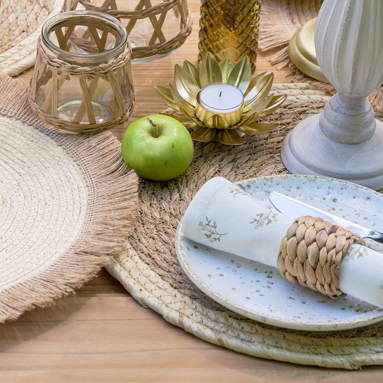 Platzset Stroh Boho Tischdeko Boho-Tischdekoration mit rundem Stroh-Platzset, Kerzenhalter, Apfel, Geschirr und Naturaccessoires auf einem gedeckten Holztisch.