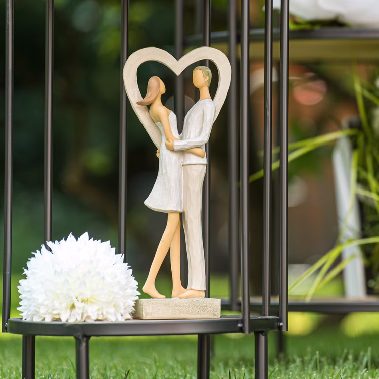 Brautpaar Figur Stehend Liebe Weiß mit Herz Tischdeko Hochzeits-Deko Tisch-Deko Deko-Ideen Geschenkidee Braut & Bräutigam Dekofigur Paar im Herzrahmen, aufgestellt draußen auf einem schwarzen Tisch mit Blume daneben.