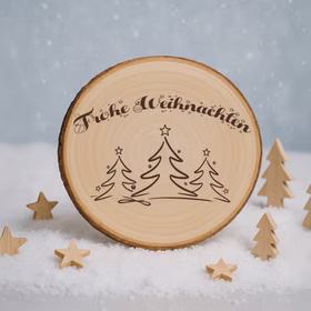 Gravierte Baumscheibe „Frohe Weihnachten“ stehend im Kunstschnee, umgeben von kleinen Holzsternen und Holztannen vor winterlicher Schneelandschaft.