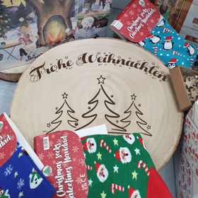 Große Baumscheibe mit Gravur, eingebettet in ein festliches Arrangement aus Tannenzweigen, goldenen Kugeln, Teelichtern und Kunstschnee, im Hintergrund warm leuchtende Bokeh-Lichter.