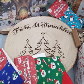 Große gravierte Baumscheibe mit „Frohe Weihnachten“ und Tannenmotiv, umgeben von Weihnachtsartikeln wie Socken und Adventskalendern auf einem Tisch.