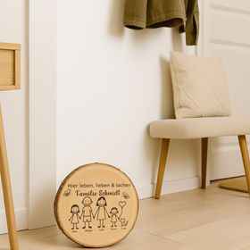 Personalisierte Holzscheibe mit Familienmotiv lehnt an einer Backsteinwand neben einer grünen Zimmerpflanze in einem natürlichen Wohnraumambiente.