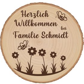 Personalisierte Baumscheibe mit Rinde und Gravur „Herzlich Willkommen bei Familie Schmidt“, dekoriert mit Schmetterlingen und Blumenmotiv, platziert in einem farbenfrohen Sommer-Blumenbeet am Hauseingang.