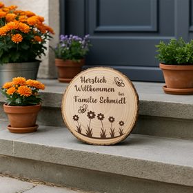 Personalisierte Baumscheibe mit Rinde und Gravur „Herzlich Willkommen bei Familie Schmidt“, dekoriert mit Schmetterlingen und Blumenmotiv, platziert in einem farbenfrohen Sommer-Blumenbeet am Hauseingang.