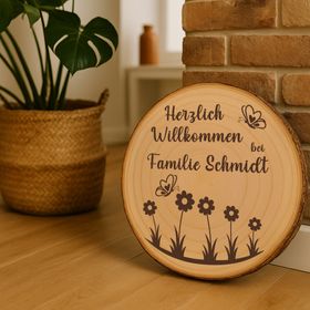 Personalisierte Baumscheibe mit Rinde und Gravur „Herzlich Willkommen bei Familie Schmidt“, dekoriert mit Schmetterlingen und Blumenmotiv, platziert in einem farbenfrohen Sommer-Blumenbeet am Hauseingang.