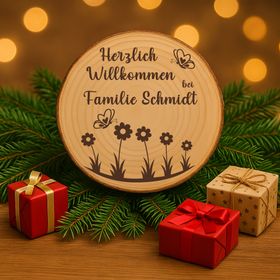 Personalisierte Baumscheibe mit Rinde und Gravur „Herzlich Willkommen bei Familie Schmidt“, dekoriert mit Schmetterlingen und Blumenmotiv, platziert in einem farbenfrohen Sommer-Blumenbeet am Hauseingang.