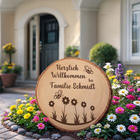 Personalisierte Baumscheibe mit Rinde und Gravur „Herzlich Willkommen bei Familie Schmidt“, dekoriert mit Schmetterlingen und Blumenmotiv, platziert in einem farbenfrohen Sommer-Blumenbeet am Hauseingang.