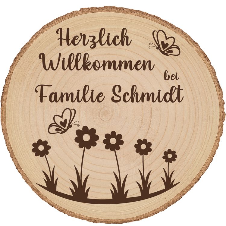 Personalisierte Baumscheibe mit Rinde und Gravur „Herzlich Willkommen bei Familie Schmidt“, dekoriert mit Schmetterlingen und Blumenmotiv, platziert in einem farbenfrohen Sommer-Blumenbeet am Hauseingang.