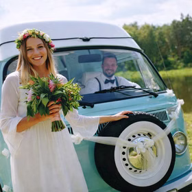Braut mit Blumenstrauß steht vor einem hellblauen Hochzeitsauto, das mit weißen Organza-Girlanden geschmückt ist.