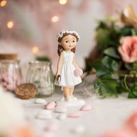 Dekofigur Mädchen zur Erstkommunion auf dekoriertem Tisch mit Blumen, Glasgefäßen und Pastellfarben im Hintergrund.