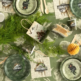 Draufsicht auf Jurassic World Partyzubehör mit Tellern, Bechern, Servietten, Geschenktüte und grüner Tischdecke.