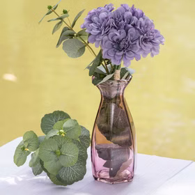 Rosafarbene Glasvase mit Blumen und grünen Blättern dekorativ inszeniert – moderne Tischdeko für Wohnräume.