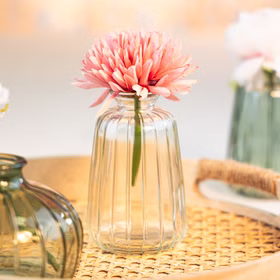 Transparente Glasvase Verone mit rosa Blüte als Tischdeko auf geflochtener Unterlage in heller, festlicher Umgebung.