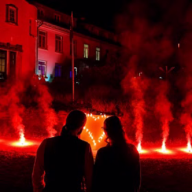 Zwei Personen betrachten ein brennendes Lichterbild mit Herz-Motvi und mit Bengalfeuern vor einem Gebäude bei Nacht.