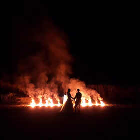 Brautpaar steht Hand in Hand vor einer Reihe brennender Bengalfeuer bei Nacht und betrachtet ein romantisches Lichterbild im Hintergrund.