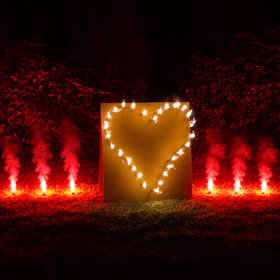 Lichterbild in Herzform mit Bengalfeuern auf einer Wiese bei Nacht.